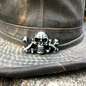 Może przedstawiać: Srebrny emblemat czaszki i skrzyżowanych piszczeli jest przymocowany do zwietrzałego, ciemnoszarego kapelusza. Emblemat jest szczegółowy z widocznymi zębami i strukturą kości. Kapelusz ma zniszczony wygląd, z widocznymi szwami i teksturą.