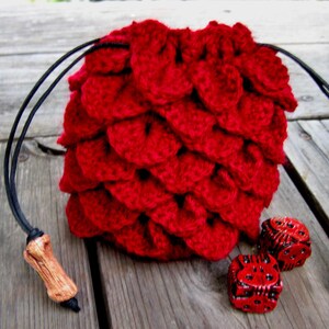 May include: Red crocheted dice bag with black drawstrings and wooden toggles. Two red dice are in front of the bag.