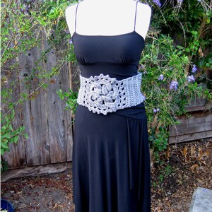 May include: A gray crocheted belt with a large floral design. The belt is worn over a black dress.