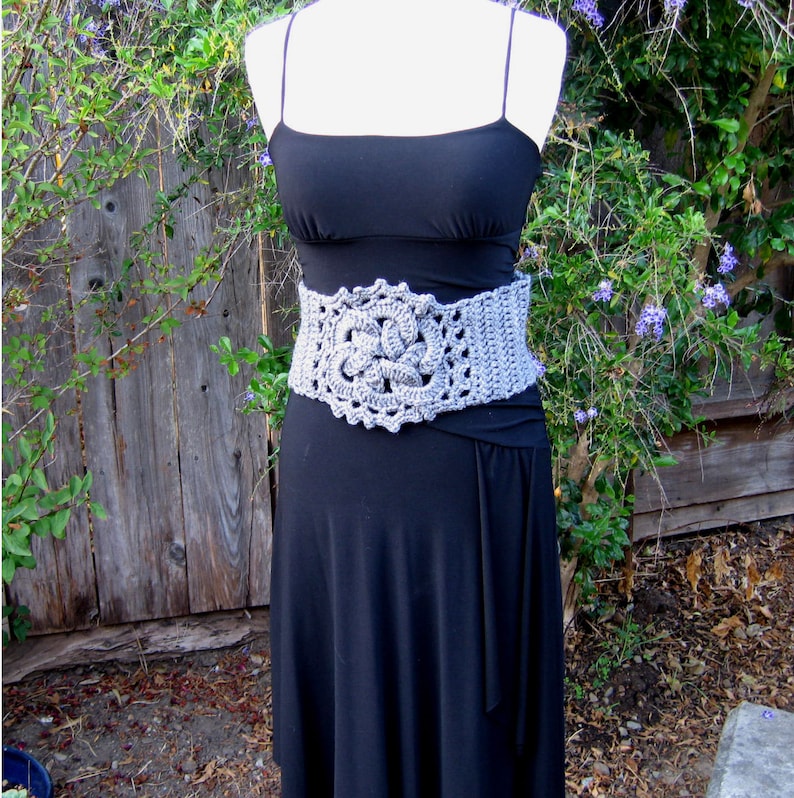 May include: A gray crocheted belt with a large floral design. The belt is worn over a black dress.