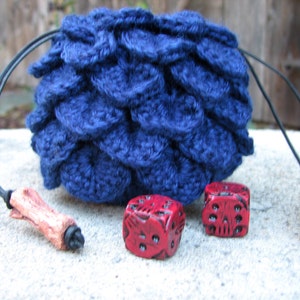 May include: A blue crocheted dice bag with a drawstring closure and wooden toggles. Two red dice are in front of the bag.