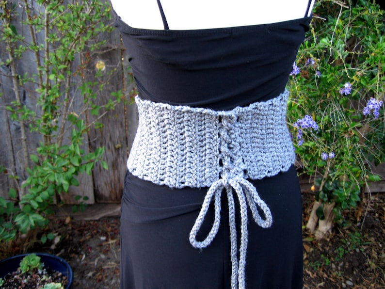 May include: A gray crocheted corset with a lace-up back. The corset is tied at the front with long gray strings.
