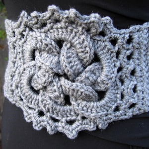 Close up of medium light grey wide crochet belt, with Celtic knot design in the middle. It appears to have a corset tie in the back. The figure is wearing a black dress and it seems to be in an outdoor  environment.