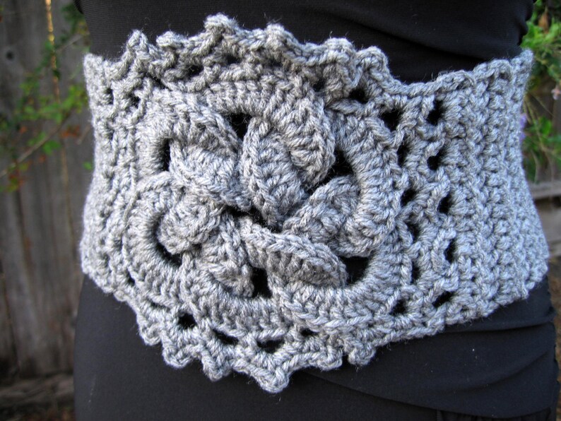 Close up of medium light grey wide crochet belt, with Celtic knot design in the middle. It appears to have a corset tie in the back. The figure is wearing a black dress and it seems to be in an outdoor  environment.
