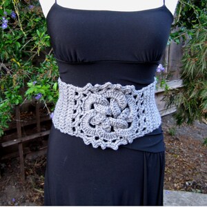 May include: A gray crocheted belt with a floral design. The belt is worn over a black dress.