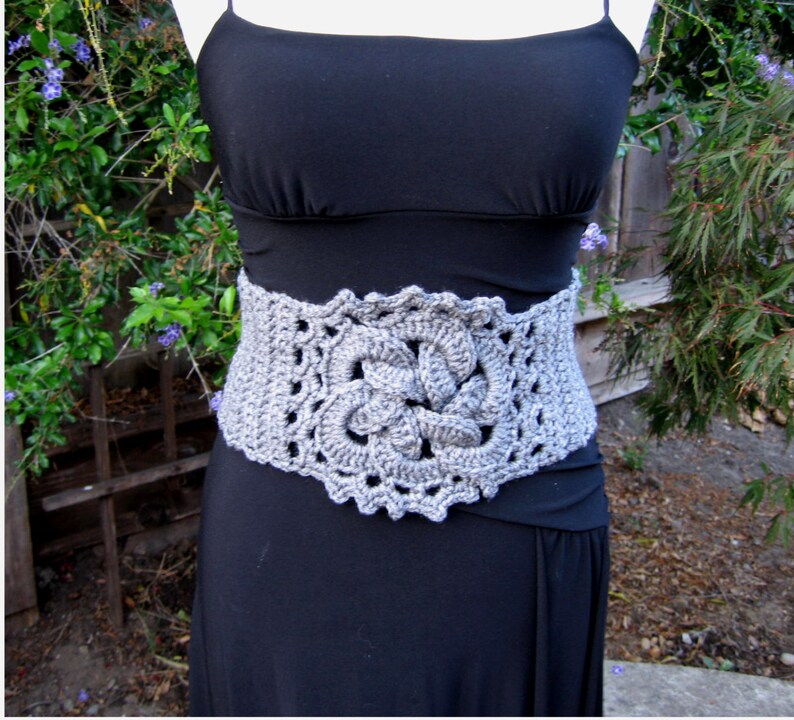 May include: A gray crocheted belt with a floral design. The belt is worn over a black dress.
