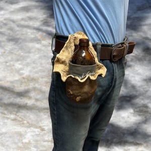 May include: A brown leather beer bottle holder attached to a brown belt. The holder is made of textured leather and has a decorative edge. A brown glass bottle is inside the holder. The person is wearing blue jeans and a blue shirt.