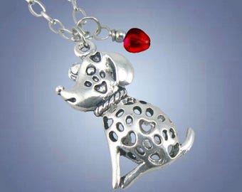 May include: A silver-toned metal necklace featuring a pendant shaped like a sitting dog with a cut-out pattern of small hearts and circles on its body. A small, smooth, bright red bead hangs from the silver chain next to the dog pendant.
