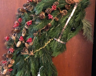 May include: A green horse head Christmas wreath decorated with pine cones, red berries, and gold garland.