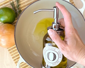 Puede incluir: Una botella de vidrio de aceite de oliva con un dispensador de bomba plateado. La botella tiene una etiqueta blanca con el texto "Olive Olive Olive Olive" y una letra "O" grande en el centro.