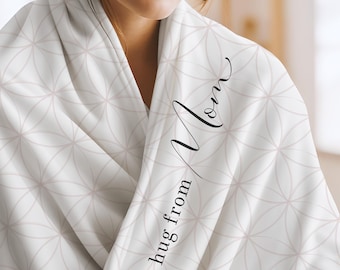 May include: A white blanket featuring a repeating geometric pattern of thin, light pink intersecting circles. The fabric is draped, and the text "a hug from Mom" is printed in a black, elegant script font vertically along the folded edge.