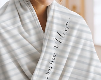 May include: A soft white blanket featuring horizontal light gray stripes. The fabric is draped over shoulders and displays the black printed text "a hug from Melissa" in a combination of elegant script and clean sans-serif typography.