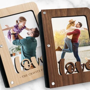 Op de afbeelding: Twee houten fotoalbums met zwarte randen en metalen accenten. Elk album heeft een uitsnijding van het woord "love" en een fotovenster. Een album is van licht hout met een foto van een vader die zijn dochter omhoog houdt. Het andere album is van donker hout met een foto van een stel dat elkaar omhelst. De tekst op het album van licht hout luidt "THE GRAVITZ FAMILY".