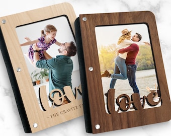 Puede incluir: Dos álbumes de fotos de madera con bordes negros y detalles metálicos. Cada álbum tiene un recorte de la palabra "love" y una ventana para fotos. Un álbum es de madera clara con una foto de un padre sosteniendo a su hija en alto. El otro álbum es de madera oscura con una foto de una pareja abrazándose. El texto en el álbum de madera clara dice "THE GRAVITZ FAMILY".