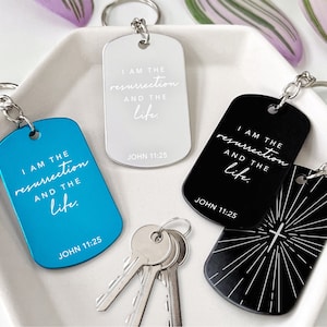 Könnte beinhalten: Schlüsselanhänger in Blau, Silber und Schwarz, jeweils mit einem rechteckigen Erkennungsmarken-Design. Die Anhänger tragen den Text "I AM THE resurrection AND THE life. JOHN 11:25" in einer handschriftlichen Schriftart. Ein schwarzer Schlüsselanhänger hat ein Kreuzdesign.