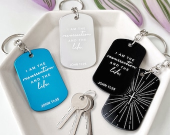 May include: Keychains in blue, silver, and black, each with a rectangular dog tag design. The tags feature the text "I AM THE resurrection AND THE life. JOHN 11:25" in a handwritten style font. One black keychain has a cross design.