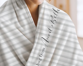 May include: A soft throw blanket featuring a light gray and white horizontal striped pattern. The fabric is draped and includes the black cursive text "a hug from Aunt Beth" printed vertically along the folded edge of the material.