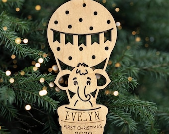 May include: A light-colored wooden ornament shaped like a hot air balloon with a cartoon elephant in the basket. The balloon features polka dots and a banner. The basket is engraved with the name EVELYN, the text FIRST CHRISTMAS, and 2029.