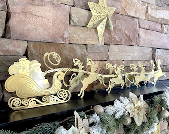 May include: Gold-colored Christmas decoration featuring Santa Claus in a sleigh pulled by reindeer. A gold star is mounted on the wall above the sleigh. The sleigh and reindeer are on a black surface, with a garland of greenery and ornaments below.