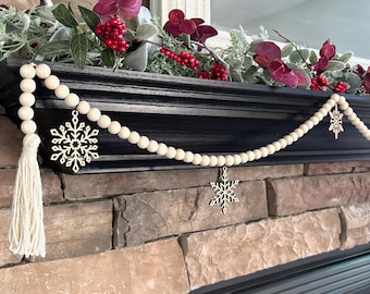 May include: A wooden bead garland with snowflake ornaments and a tassel. The garland is draped over a black mantel, decorated with faux greenery and red berries. A decorative accent for the holiday season.