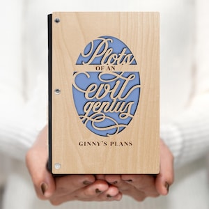 May include: A wooden notebook with a blue oval cutout that says "Plots of an evil genius" and "Ginny's Plans" on the front.