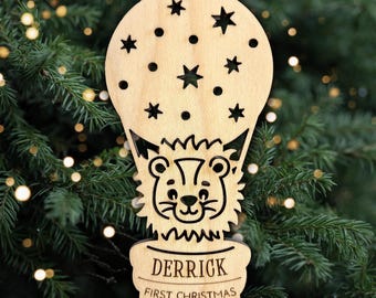 May include: A light-colored wooden ornament shaped like a hot air balloon featuring a cutout star pattern and a central lion illustration. The basket section is engraved with the name DERRICK, the text FIRST CHRISTMAS, and the year 2029.