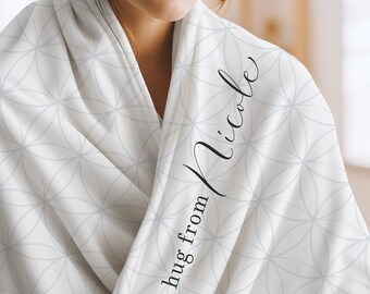 May include: A white blanket featuring a repeating light gray geometric flower of life pattern. The fabric is draped and displays the black cursive text "a hug from Nicole" printed vertically along the folded edge of the material.