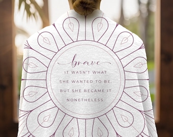 May include: White throw blanket with a mandala design in a light purple color. The center of the mandala features a circle with the text "brave" in cursive script. The text below reads "It wasn't what she wanted to be. But she became it nonetheless."
