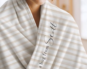 May include: A soft throw blanket featuring a pattern of horizontal white and beige stripes. The fabric is draped over shoulders and displays the black cursive text "a hug from Scott" printed vertically along the folded edge of the material.