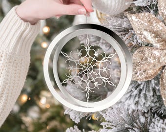 May include: A silver snowflake ornament with intricate cut-out details is held up against a decorated Christmas tree. The ornament is circular with a layered design, and the snowflake is the focal point. The tree has white and gold accents.