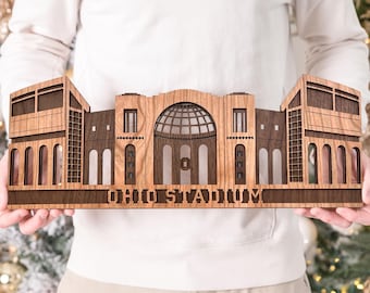 Może przedstawiać: Drewniana replika stadionu Ohio, z misternymi detalami architektonicznymi. Model stadionu wykonany jest z jasnego i ciemnego drewna, z wyciętym napisem "OHIO STADIUM" w podstawie. Dekoracyjny przedmiot dla fanów sportu.