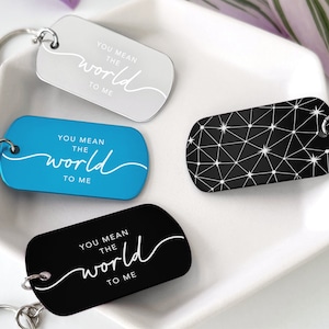 以下が含まれることがあります： 異なるデザインのキーホルダーが3つあります。1つは銀色で「You mean the world to me」というテキストが書かれています。もう1つは青色で同じテキストが書かれています。3つ目は黒色で白い幾何学模様が描かれています。