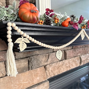 May include: A wooden bead garland with a white tassel and wooden leaf accents. The garland is draped over a black mantel with fall decor, including a pumpkin, gourds, and pine cones.
