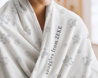 May include: A white blanket featuring a repeating pattern of light gray paw prints. The fabric is draped and includes the black text "snuggles from ZEKE" printed vertically along the front fold of the material, creating a soft and textured appearance.