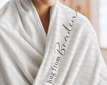 May include: A white blanket featuring a repeating beige geometric pattern of interconnected rounded shapes. The fabric displays the black cursive text "a hug from Braden" printed vertically along the folded edge of the material.