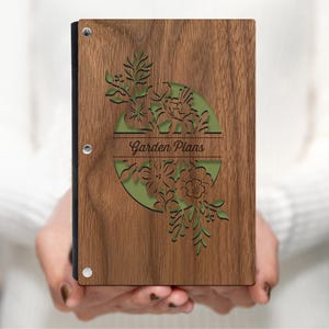 Op de afbeelding: Een houten map met een groen bloemmotief en de tekst "Garden Plans" in een cirkel. De map is gemaakt van walnoot hout en heeft een zwarte rug.