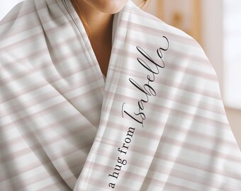 May include: A soft throw blanket featuring a pattern of horizontal light pink and white stripes. The fabric is draped and includes the black cursive text "a hug from Isabella" printed vertically along the folded edge of the material.