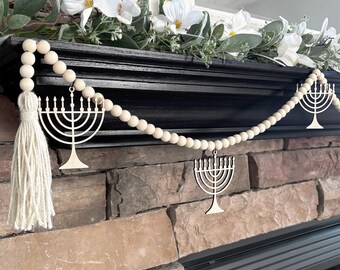 May include: A decorative garland featuring natural light-colored wooden beads strung together, accented with three flat wooden menorah cutouts and a long, cream-colored fiber tassel at one end, designed for hanging display across a horizontal surface.