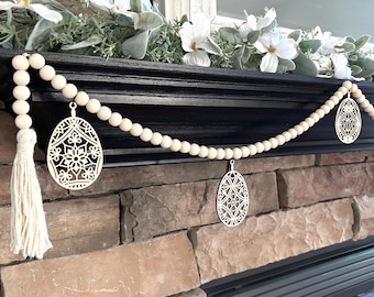 May include: A decorative Easter garland featuring wooden beads and laser-cut egg ornaments. The garland includes a white tassel and is draped over a black mantel, with faux floral accents. The egg ornaments have intricate cut-out designs.