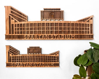 Könnte beinhalten: Zwei Wandkunstwerke aus Holz des Spartan Stadiums. Das größere Stück befindet sich über dem kleineren. Beide bestehen aus hellem und dunklem braunem Holz, wobei die Worte "SPARTAN STADIUM" in das Design geschnitten sind. Das Stadiondesign enthält ein Spartan-Logo.