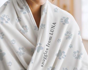 May include: A white fleece blanket featuring a repeating pattern of light gray paw prints. The fabric is draped and includes the black text "snuggles from LUNA" printed vertically along the folded edge of the soft material.