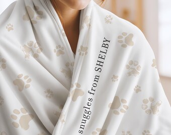 May include: A white blanket featuring a repeating pattern of light beige paw prints. The fabric is draped and includes the black printed text "snuggles from SHELBY" running vertically along the folded edge of the material.