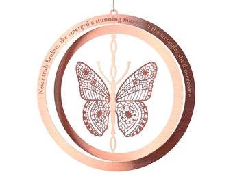 May include: A rose gold metal ornament featuring a detailed butterfly design. The butterfly is centered within two concentric circles, with the text "Never truly broken, she emerged a stunning mosaic of the struggles she'd overcome." The ornament hangs from a silver chain.