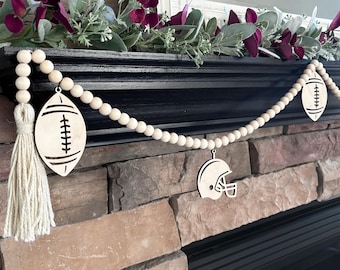 May include: A decorative garland featuring natural light-colored wooden beads strung together, accented with a cream-colored yarn tassel at one end. The garland includes three wooden cutouts: two football shapes and one football helmet shape.