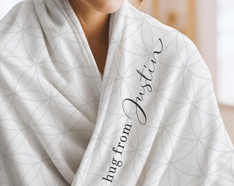 May include: A white blanket featuring a repeating light gray geometric flower of life pattern. The fabric is draped over shoulders and displays the black cursive text a hug from Justin printed vertically along the folded edge of the material.
