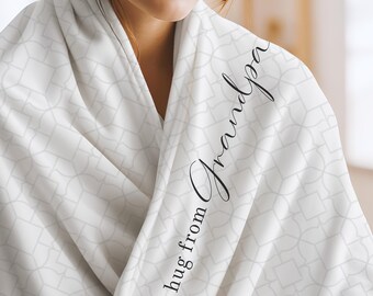 May include: A white blanket featuring a repeating light gray geometric pattern of squares and curved lines. The fabric is draped over shoulders and displays the black cursive text "a hug from Grandpa" printed vertically along the folded edge.