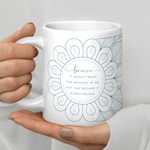 May include: White ceramic mug with a blue and white mandala design. The text "brave" is written in cursive inside the mandala. The text below the mandala reads "It wasn't what she wanted to be. But she became it nonetheless."