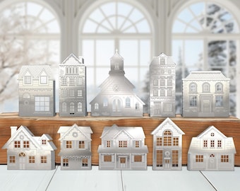 May include: A collection of silver-colored metal house and church figurines. The detailed miniature buildings include various architectural styles, with windows and doors cut out. They are arranged on a wooden surface against a wintery backdrop.
