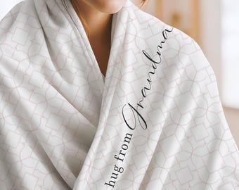May include: A white throw blanket featuring a repeating geometric pattern in soft pink lines. The fabric is draped over shoulders and displays the black cursive text "a hug from Grandma" printed vertically along the folded edge of the material.