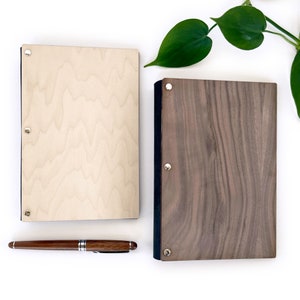 May include: Two wooden notebooks with black covers and silver rivets. The notebook on the left has a light wood grain pattern, while the notebook on the right has a dark wood grain pattern.